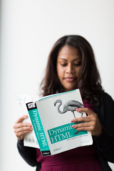 A woman with medium skin tone and long, dark brown hair is standing indoors against a plain white background, holding an open book titled 'Dynamic HTML' published by O'Reilly. She appears to be examin
